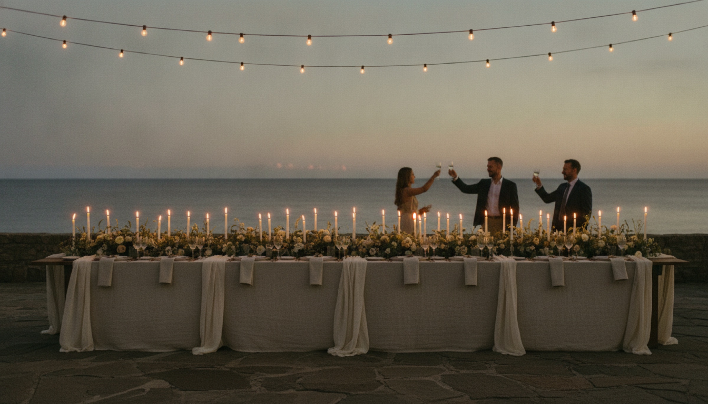 Long-table wedding dinner on a coastal terrace at dusk, string lights overhead